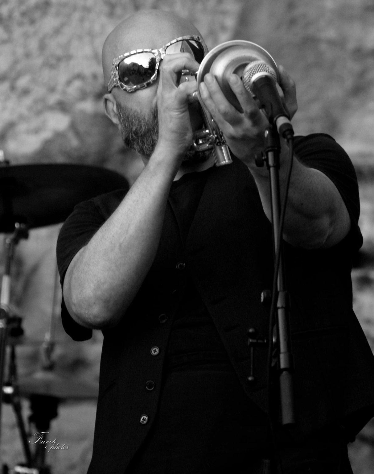 une photo en noir et blanc de Vinz, ses grosses lunettes noires, sa trompette avec une sourdine derrière un micro sur pied